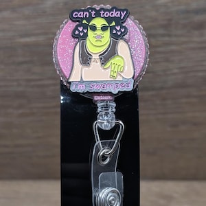 May include: A badge reel with a cartoon character wearing sunglasses and the text "can't today, I'm swamped." The reel has a pink glitter border and a clear plastic clip.