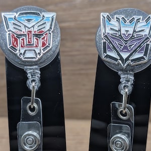 May include: Two retractable badge holders with a Transformers logo. One features a red, white, and blue design, while the other has a purple, blue, and white color scheme. Both have a silver glittery background and a clear plastic strap.