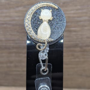 May include: A circular badge reel featuring a black glitter background with a gold-toned crescent moon adorned with clear crystals. A smooth, cream-coloured cat figure sits in front of the moon. The badge reel has a clear plastic strap and a metal clip.