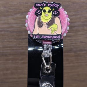 May include: A badge reel featuring a circular pink glitter base with a white beaded border. Centered is a black enamel pin of a green character wearing sunglasses, with the text 'can't today' above and 'i'm swamped' below in pink lettering.