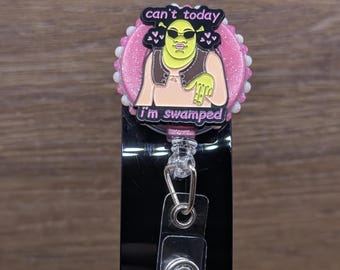 May include: A badge reel featuring a circular pink glitter base with a white beaded border. Centered is a black enamel pin of a green character wearing sunglasses, with the text 'can't today' above and 'i'm swamped' below in pink lettering.