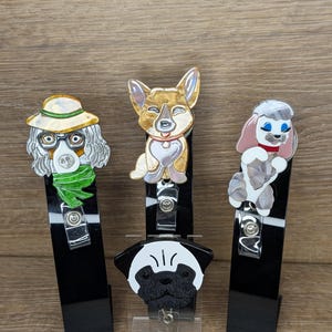 May include: Three dog-themed retractable badge holders. The designs include a poodle, a corgi, and a pug. Each badge holder has a black base with a metal clip and a clear plastic snap. The poodle has a pink bow.