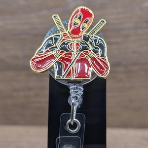 May include: A retractable badge reel with a Deadpool design. The badge reel is red, black, and gold. The Deadpool character is in a heroic pose with two swords crossed behind him. The reel has a clear plastic strap and a metal clip.