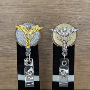 May include: Two medical symbol badge reels. One reel has a gold-coloured symbol on a silver glitter background, the other has a silver-coloured symbol on a gold glitter background. Both have a retractable cord and a metal clip.