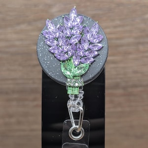 May include: A circular badge reel featuring a bouquet of purple marquise-cut crystals arranged as lavender flowers, with green crystal leaves and a clear crystal-encrusted stem base, all set against a dark grey, glitter-flecked circular background.