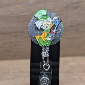 May include: A badge reel featuring the Mad Hatter from Alice in Wonderland. The design shows the Mad Hatter in a green hat and yellow coat, pouring tea. The reel has a glittery, grey background and a clear plastic clip. The badge reel is attached to a black plastic holder.