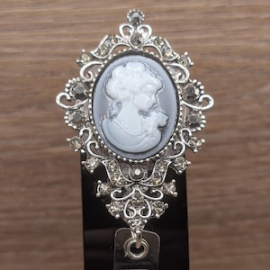 May include: A silver-toned badge reel featuring an oval cameo with a white profile portrait on a dark grey background. The frame is ornate, decorated with clear rhinestones in a filigree pattern, extending into a decorative base with a metal loop.