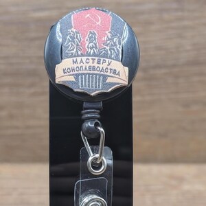May include: A black retractable badge reel with a decorative emblem. The emblem features a red background with a hammer and sickle, and the text "MASTER OF HEMP FARMING" in a banner. The reel has a metal clip and a clear plastic strap.