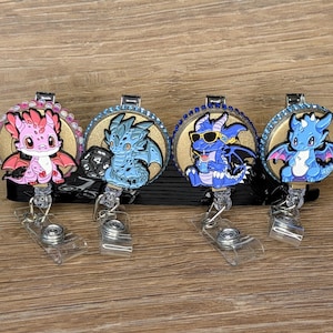 May include: Four dragon-themed badge reels. Each reel features a different dragon design, including a pink, blue, and purple dragon. The reels have a clear plastic clip and a retractable cord.