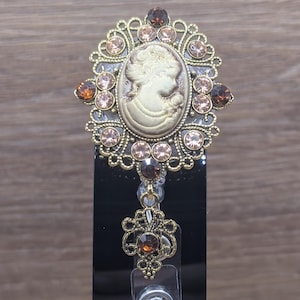 May include: A gold-toned badge reel featuring an oval cameo centrepiece with a profile portrait of a person. The frame is decorated with light pink and dark amber rhinestones in a filigree setting, with a dangling filigree charm at the bottom.