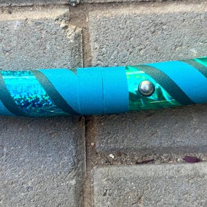 May include: A section of a teal and green hula hoop. The hoop features teal tape and dark teal diagonal stripes. A silver metal button secures the tape. The hoop is on a grey brick surface.