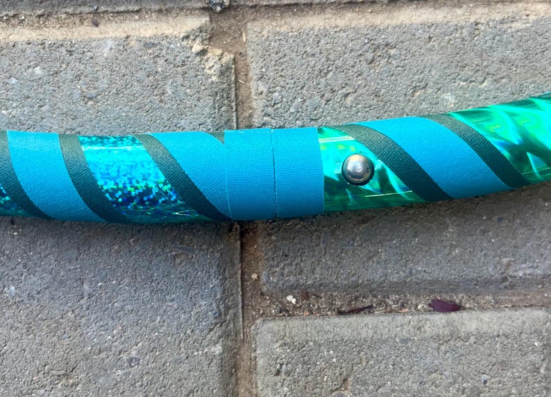 May include: A section of a teal and green hula hoop. The hoop features teal tape and dark teal diagonal stripes. A silver metal button secures the tape. The hoop is on a grey brick surface.