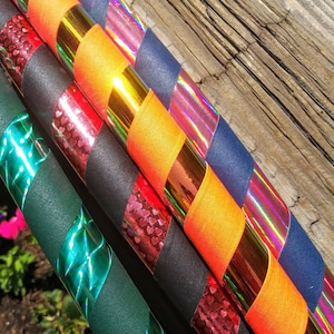May include: Close-up of several colourful hula hoops. The hoops are decorated with a variety of tapes in shades of orange, red, green, black, and blue. Some tapes have a shiny, metallic finish, while others have patterns.