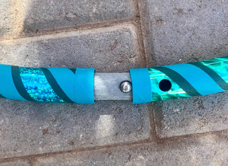 May include: A section of a teal and green hula hoop, with a connecting mechanism. The hoop has a teal and green patterned design with teal bands. The connection point is a silver metal piece with a silver button.