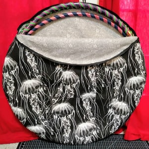 May include: A circular black bag with a zip, decorated with a white jellyfish design. The bag is open, showing colourful hula hoops inside. The interior lining has a light grey pattern. The bag is designed for carrying hula hoops.