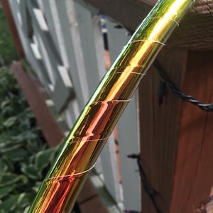 Puede incluir: Primer plano de un aro dorado iridiscente con un patrón en espiral. El aro está hecho de un material flexible y está envuelto en una película brillante e iridiscente.