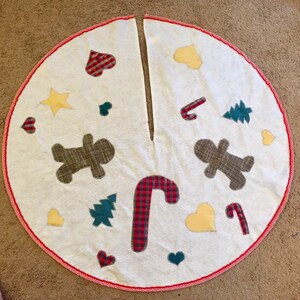 May include: White Christmas tree skirt with red trim. The skirt is decorated with gingerbread men, candy canes, hearts, stars, and Christmas trees.