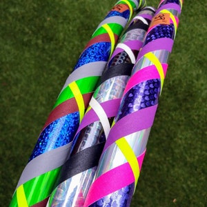 May include: Three decorated hula hoops. One is blue glitter, another has purple hearts, and the third has a mix of black, silver, and purple. Each hoop is adorned with stripes of neon green, yellow, and pink tape. The hoops are set against a green background.