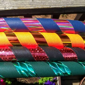 May include: A stack of colourful hula hoops. Each hoop features a combination of solid-coloured fabric and iridescent tape in various colours, including blue, yellow, red, black, and green. The hoops are stacked on top of each other.