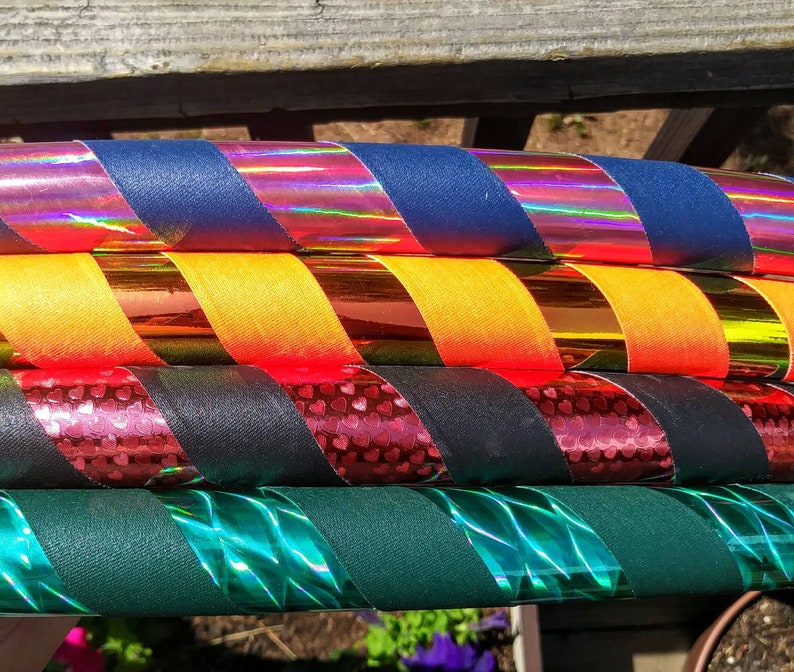 May include: A stack of colourful hula hoops. Each hoop features a combination of solid-coloured fabric and iridescent tape in various colours, including blue, yellow, red, black, and green. The hoops are stacked on top of each other.