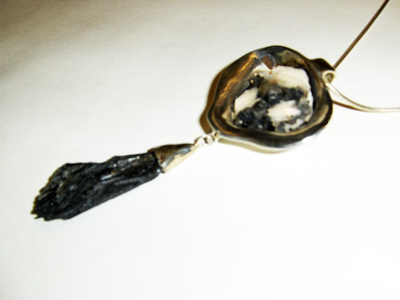 Puede incluir: Un collar de plata con un colgante de piedra negra. El colgante es una piedra negra larga y delgada con un engaste de plata. El collar est&aacute; sobre un fondo blanco.