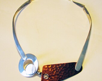 Puede incluir: Un collar de plata con un colgante de cobre martillado. El colgante tiene forma triangular con esquinas redondeadas y tiene una textura martillada. El collar está hecho de dos bandas de plata que están conectadas por un anillo de plata.