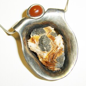 May include: A silver pendant with a carved out section that holds a brown and white crystal. The pendant is attached to a silver chain and has a red carnelian stone at the top.