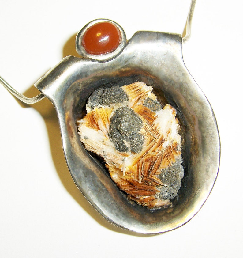May include: A silver pendant with a carved out section that holds a brown and white crystal. The pendant is attached to a silver chain and has a red carnelian stone at the top.