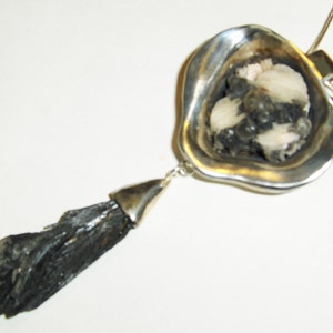 Puede incluir: Un colgante de plata con una piedra negra y blanca engastada en un marco de plata en forma de coraz&oacute;n. Una piedra negra, larga, fina y cristalina cuelga de la parte inferior del colgante.