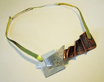 Puede incluir: Un collar de cadena dorada con un colgante geométrico hecho de plata, cobre y latón. El colgante es una combinación de una forma cuadrada y triangular.