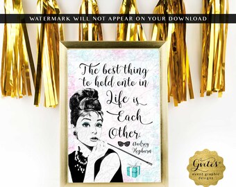 May include: A rectangular gold-framed sign featuring a black and white illustration of Audrey Hepburn with a cigarette holder. The text reads: The best thing to hold onto in life is each other. Audrey Hepburn. Includes a small blue gift box icon.