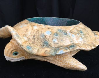 May include: A ceramic turtle-shaped planter with a textured, tan shell and a turquoise interior. The turtle has detailed features, including a patterned shell, flipper-like legs, and a detailed head with blue eyes. The planter is designed to hold plants or other decorative items.