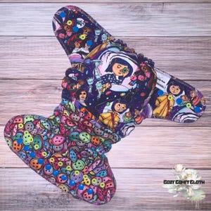 May include: Cloth diaper featuring a colorful button pattern and a dark blue pattern with characters from a movie. The diaper has a contoured shape with snaps and a soft, absorbent interior. The diaper is designed for babies and toddlers.