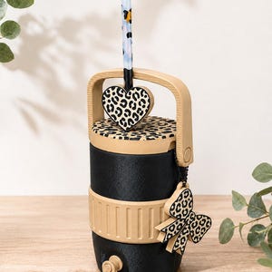 Puede incluir: Un enfriador de bebidas cilíndrico negro con asa, tapa y banda base de color beige. La tapa presenta un estampado de leopardo, un dije a juego en forma de corazón y una pajita. Un lazo con estampado de leopardo cuelga del asa y hay un pequeño grifo en la base.