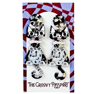 May include: A pair of black and white cat-shaped earrings with a clock design on the body. The earrings have a glittery, iridescent finish and are attached to a white card with the text "The Groovy Passport".
