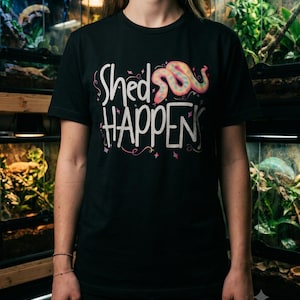 May include: A black short-sleeved t-shirt featuring the white text "Shed HAPPENS" across the chest. Above the text is a colourful, stylised snake illustration in shades of pink, blue, and yellow, accented by small decorative star shapes.