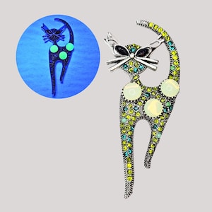 May include: A silver-toned cat brooch with a playful design. The cat is adorned with green, yellow, and blue rhinestones, with three large, round, opalescent stones. The cat has a black bow and whiskers. A smaller, glowing version of the brooch is in a blue circle.