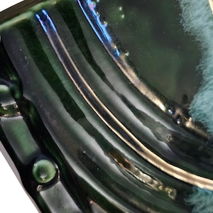 May include: A close-up view of a dark green ceramic surface with a glossy, reflective glaze. The piece features raised, curved ridges and a smooth, undulating texture. A patch of soft, light blue, fuzzy material is visible on the right side.