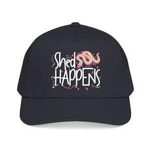 May include: A dark charcoal baseball cap featuring a graphic print with the white text "Shed HAPPENS" and a colorful, patterned snake illustration. The design is accented with small pink and teal star and squiggle shapes surrounding the text.