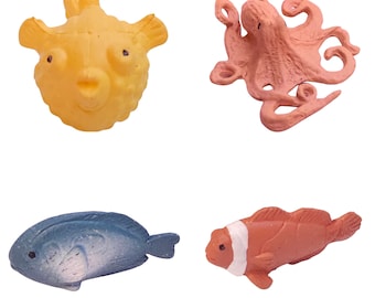 May include: A set of four small sea creature figurines including a yellow pufferfish, a pink octopus, a blue oval-shaped fish, and an orange and white striped fish. Each figurine features distinct textures and colors representing marine life.