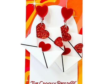 May include: Red heart-shaped earrings with glitter accents dangle from white envelope-shaped charms. The earrings are displayed on a card with a retro design and the text "THE GROOVY PASSPORT".