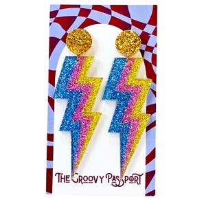 May include: Glittery lightning bolt earrings in blue, pink, and yellow with gold glitter studs. The earrings are on a white card with a red and white checkered pattern and the text "The Groovy Passport".
