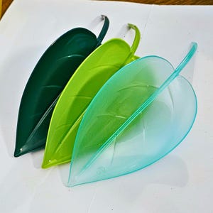 May include: Three leaf-shaped dishes arranged in a row, featuring a dark forest green opaque dish, a bright lime green opaque dish, and a translucent light turquoise blue dish. Each dish has a raised central vein and subtle leaf vein details.