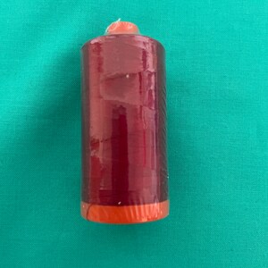 Puede incluir: Una bobina de hilo rojo con la parte superior naranja. El hilo está envuelto en plástico transparente.