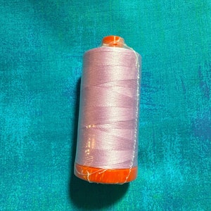 May include: A spool of lavender-colored thread wrapped in clear plastic. The spool has an orange base and top. The background is a textured teal fabric.