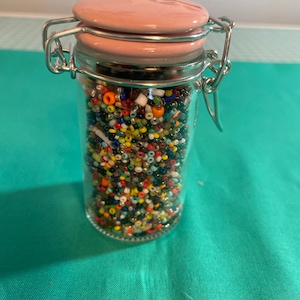 May include: A glass jar with a pink ceramic lid and a silver latch. The jar is filled with colorful seed beads.