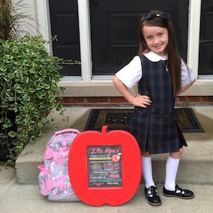 May include: A young girl in a plaid jumper and white shirt stands next to a red apple-shaped sign that reads "Ella Rose's First Day of Kindergarten." A pink backpack with bows sits nearby. The girl is wearing black shoes and white socks.