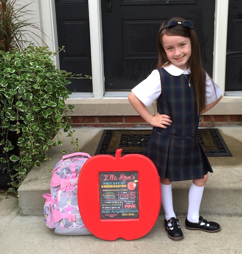 May include: A young girl in a plaid jumper and white shirt stands next to a red apple-shaped sign that reads "Ella Rose's First Day of Kindergarten." A pink backpack with bows sits nearby. The girl is wearing black shoes and white socks.