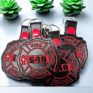May include: A set of five black leather keychains with red embroidered details. Each keychain features a firefighter's emblem with a crossed axe and hose, the words "FIRE" and "RESCUE", and a number. One keychain has the number "648" embroidered on it.