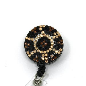 May include: A black retractable badge holder with a clear plastic strap. The badge holder has a round, black, rhinestone-covered face with a geometric pattern in gold and brown rhinestones.